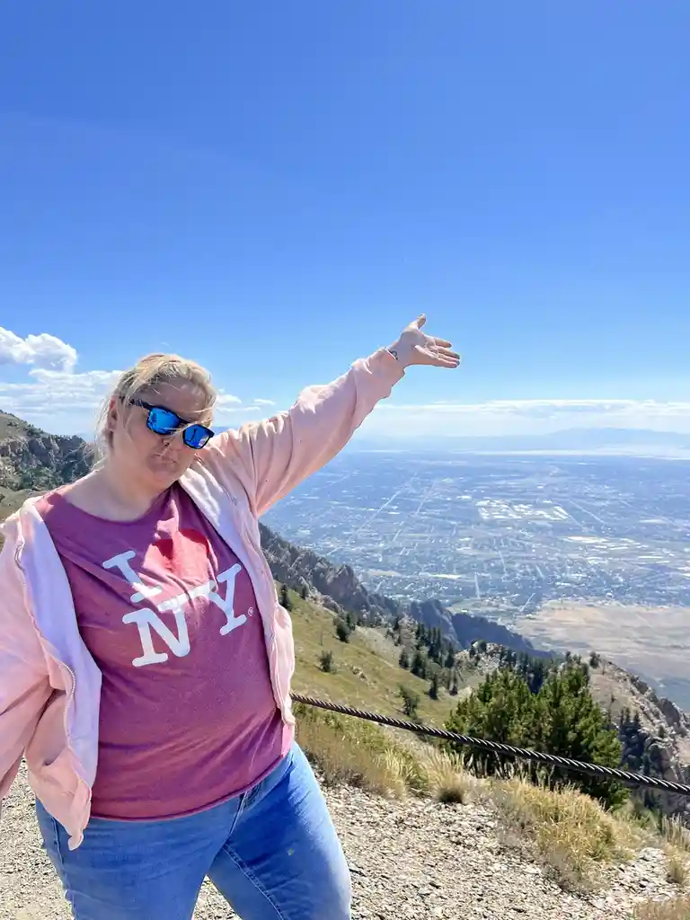 A woman wearing a pink jacket and a maroon t-shirt with 'I ❤️ NY' printed on it stands on a mountain overlook, gesturing…