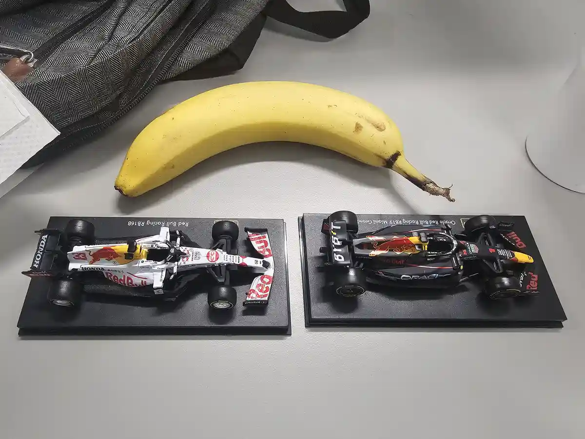 Two Red Bull Formula 1 car model toys sit side by side on black display stands, with a ripe yellow banana placed above them…