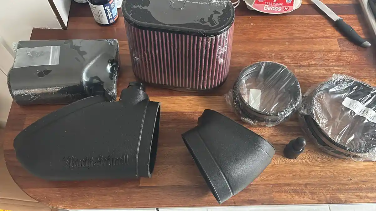 A wooden table holds various car air filter components, including a red pleated filter, black plastic housing parts, and…
