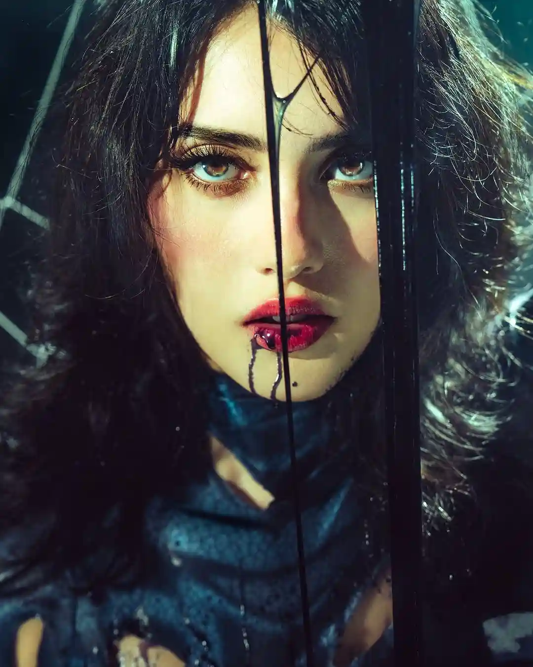 A woman with dark hair and red lipstick stares intensely through a rain-splattered window, with dark liquid dripping from…