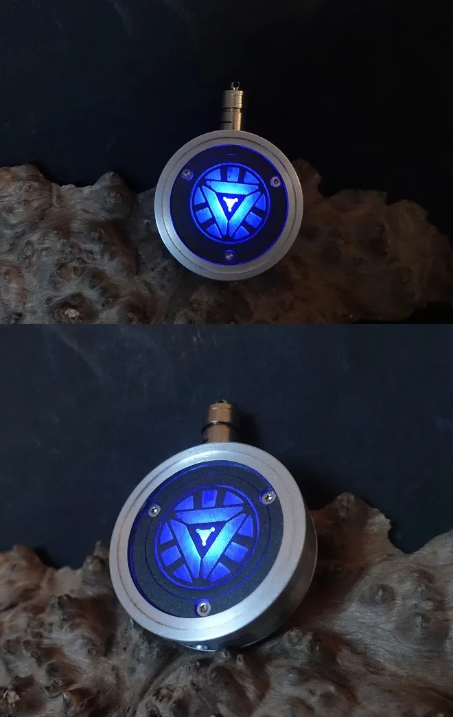 The image shows a circular, metallic device with a glowing blue arc reactor symbol at its center, resting on a piece of…