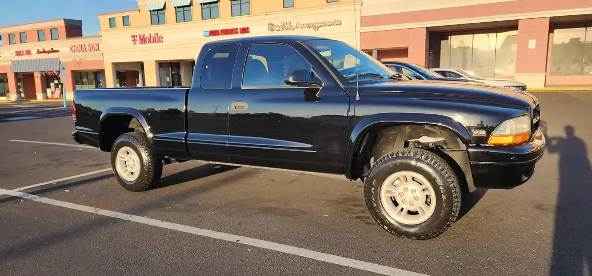 A black Dodge Ram pickup truck is parked in a commercial parking lot, its front right wheel missing and replaced with a…