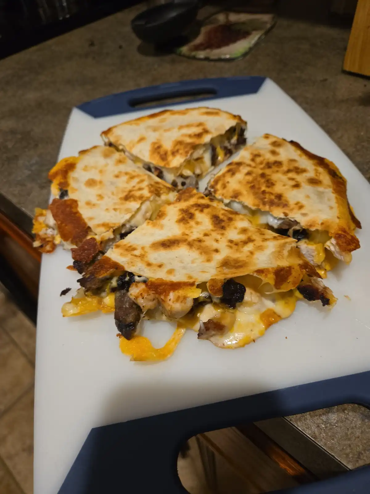 A golden-brown, sliced quesadilla with melted cheese and meat filling sits on a white cutting board with blue handles…