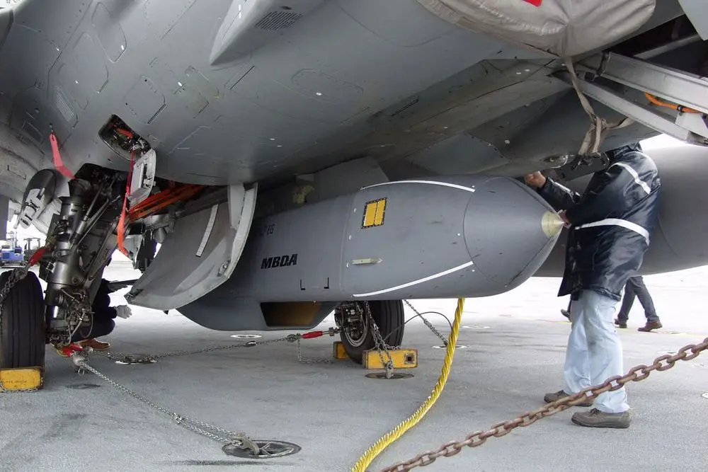 A military technician in a dark jacket and light pants is securing a large gray MBDA missile under the wing of a fighter jet…