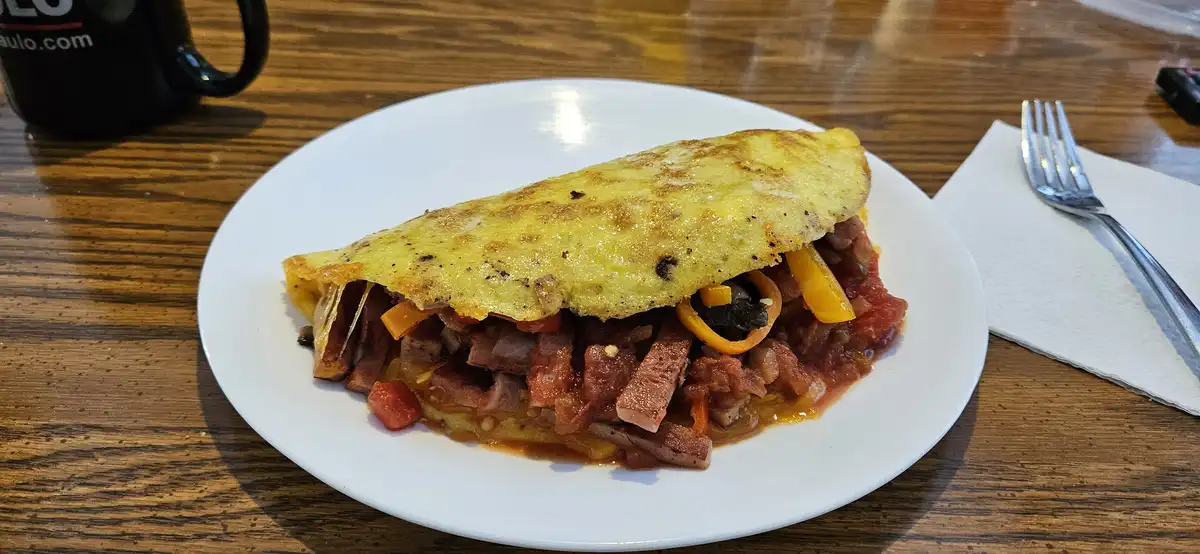 A close-up photo shows a large, folded omelette on a white plate, revealing a filling of meat, peppers, and sauce. The plate…