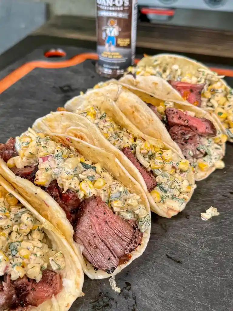 A row of four soft tacos filled with sliced steak and creamy corn salsa sit on a black cutting board. In the background, a…