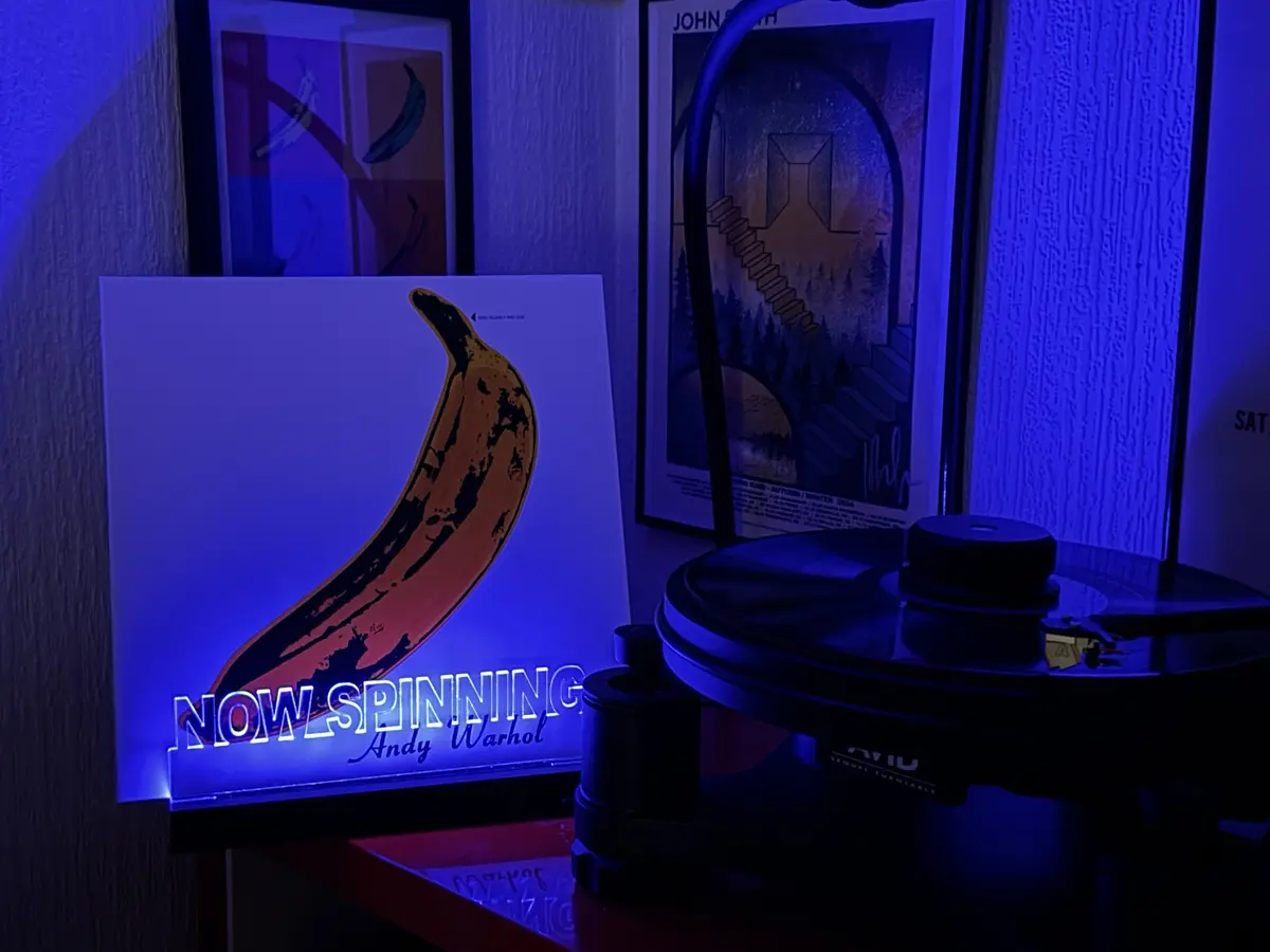 A vinyl record player sits under blue light next to a glowing sign featuring Andy Warhol's iconic banana artwork. The sign…