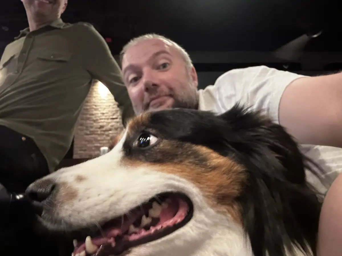 A selfie shows a man with a beard and white shirt holding a camera, with a tricolor dog's face in the foreground. The dog…