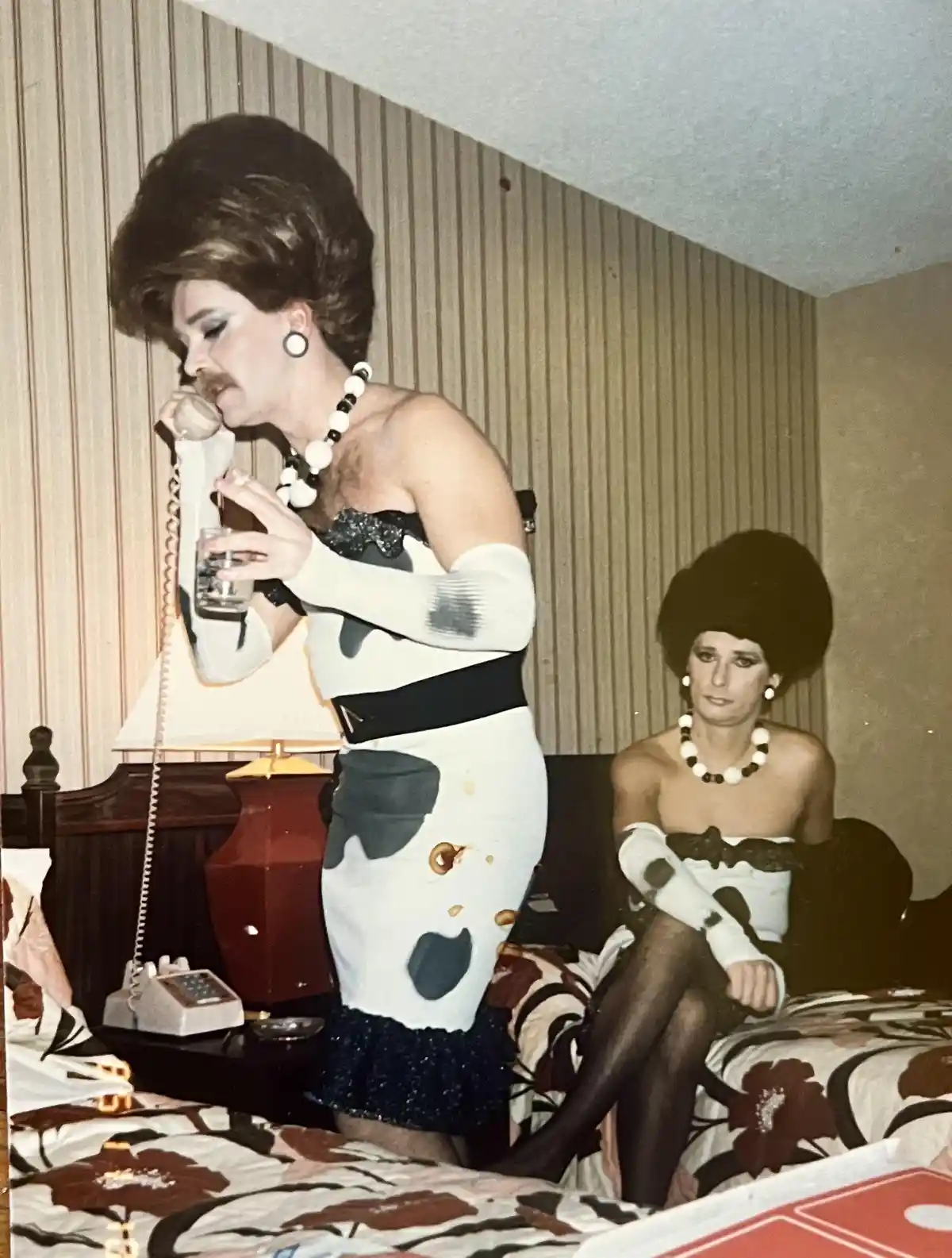 Two people in a bedroom wearing matching cow-print outfits, one standing and talking on a rotary phone while holding a…