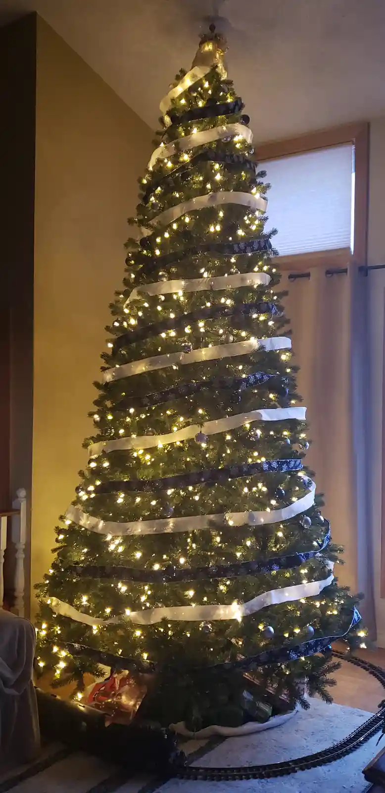 A tall, decorated Christmas tree stands indoors, adorned with alternating wide black and white ribbons spiraling around its…