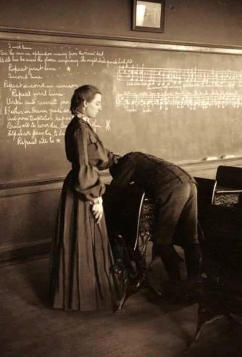 A sepia-toned historical photo shows a woman in a long dress standing beside a man in a suit who is bent over a piano. The…