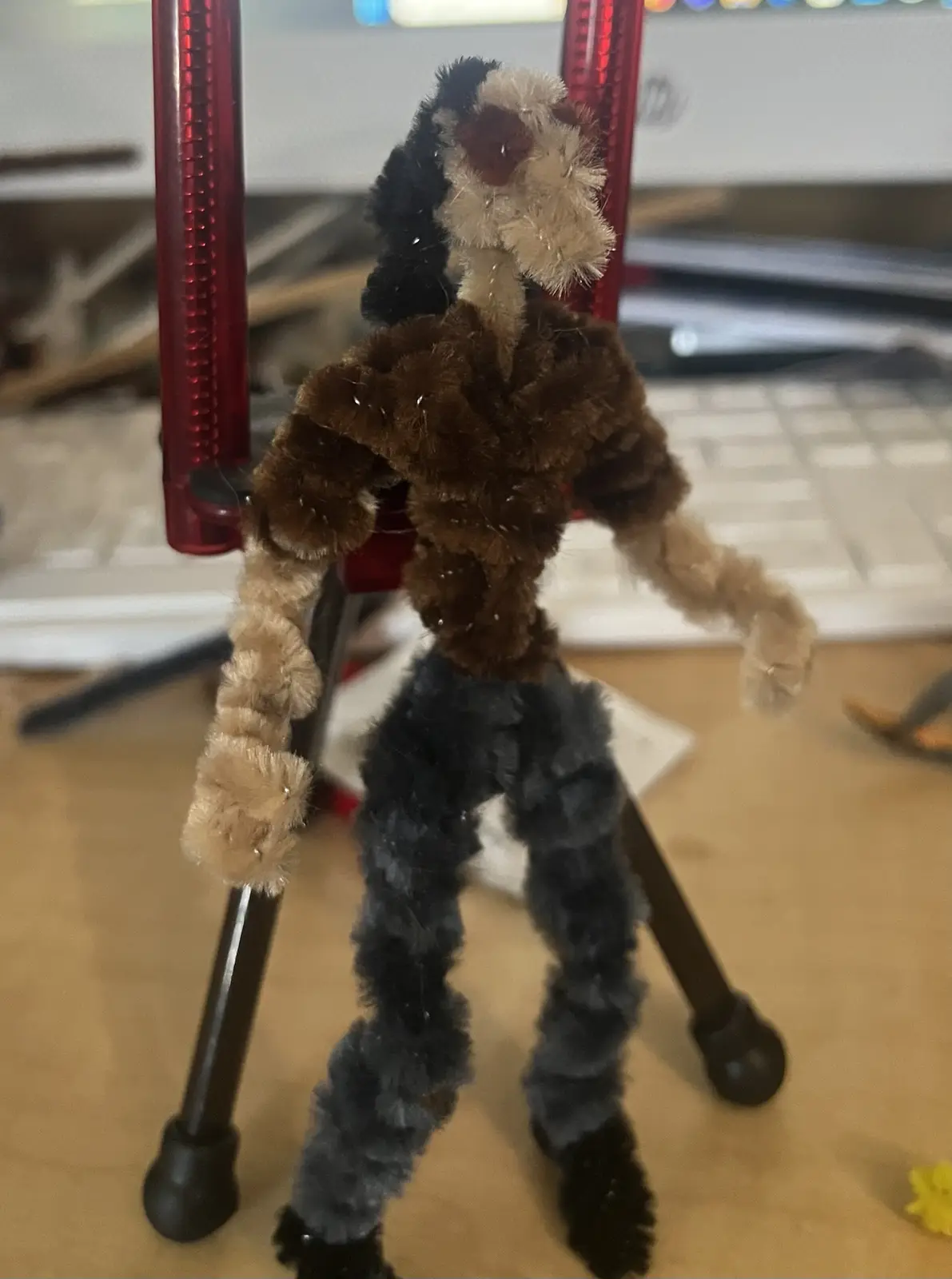 A handmade figurine made of fuzzy pipe cleaners, resembling a humanoid with a brown torso, black pants, and a…