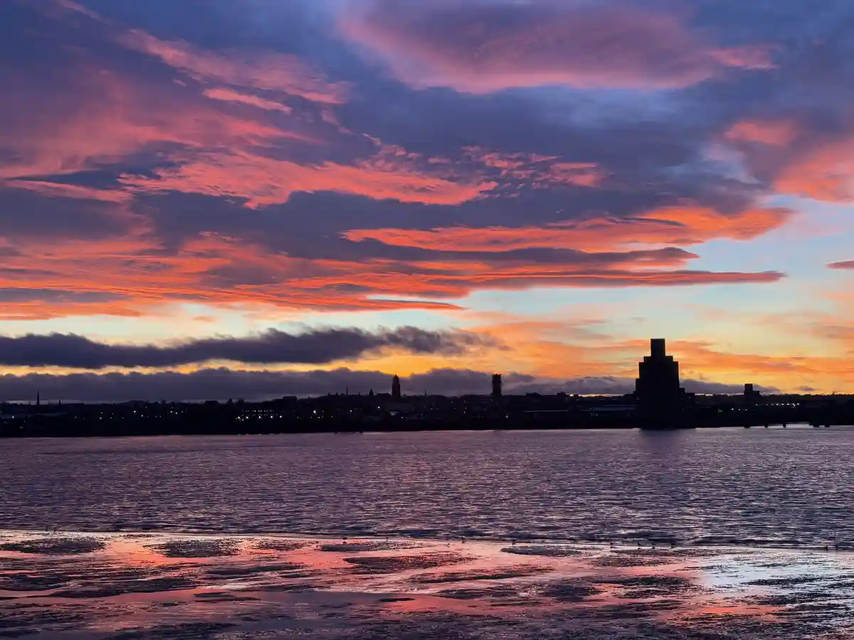 A scenic photo of a vibrant sunset over a body of water with a city skyline in silhouette, featuring dramatic pink, orange…
