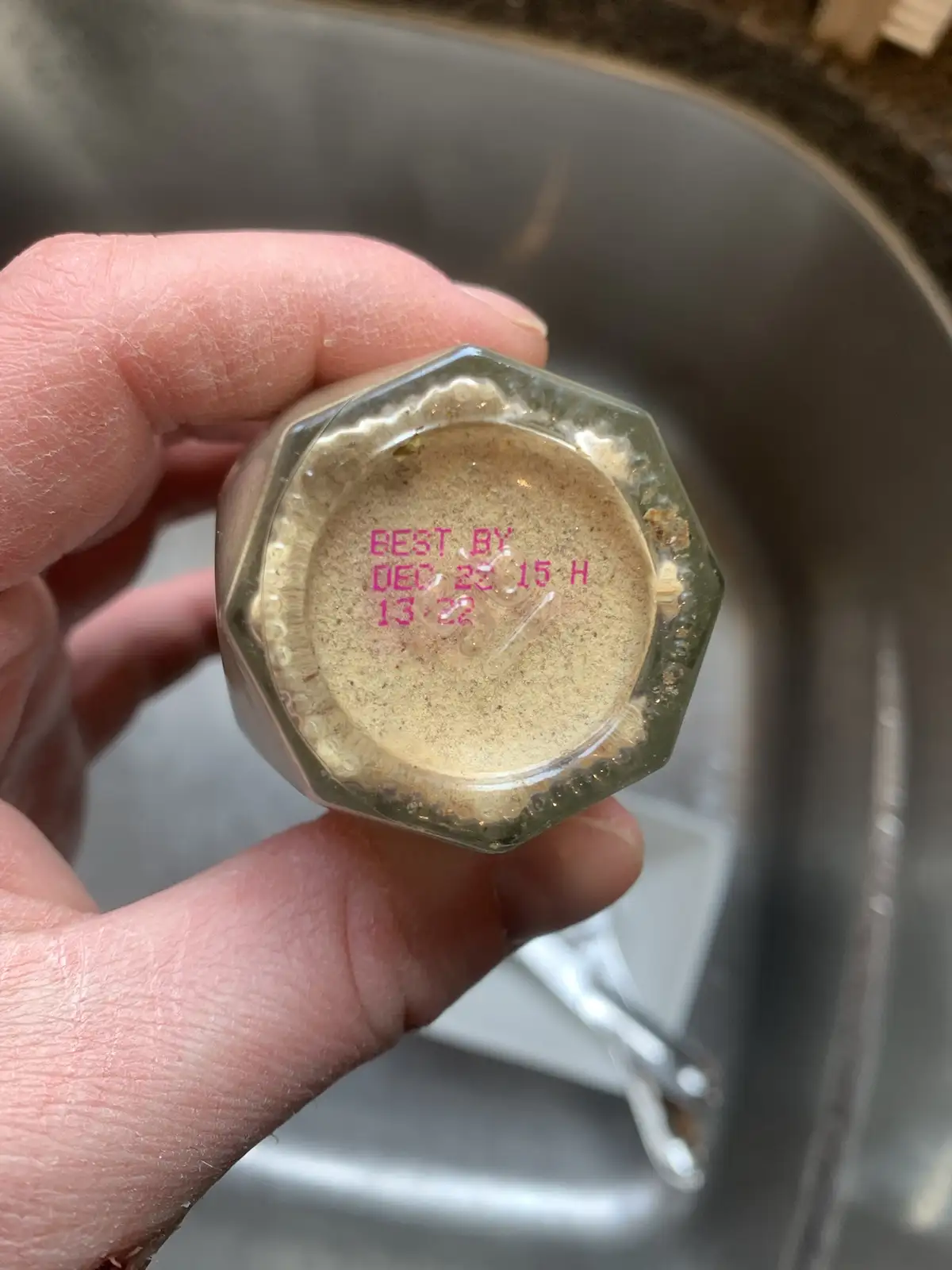 A hand holds a hexagonal glass spice jar upside down over a sink, revealing a pink 'BEST BY DEC 22 15 H 13-22' date stamp on…