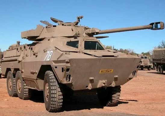 A military armored vehicle, likely an Australian Bushmaster, is shown parked on red dirt under a clear blue sky. The vehicle…