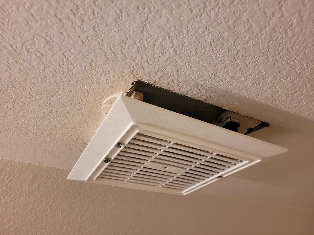 A close-up photo shows a white square air vent partially installed in a textured ceiling, with its frame hanging loosely and…