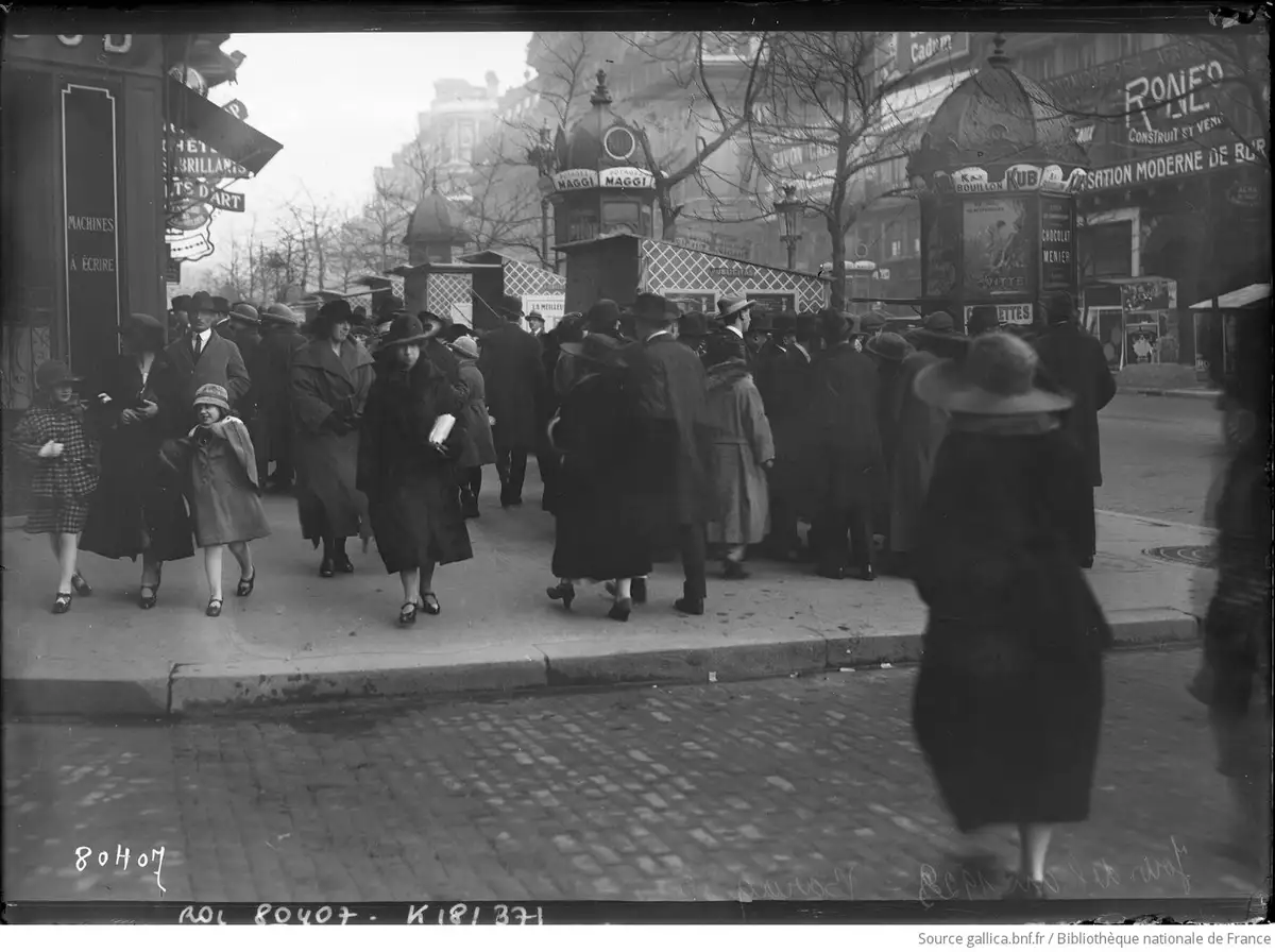 A vintage black-and-white photograph shows a crowded Parisian street scene from the early 20th century, with people in…