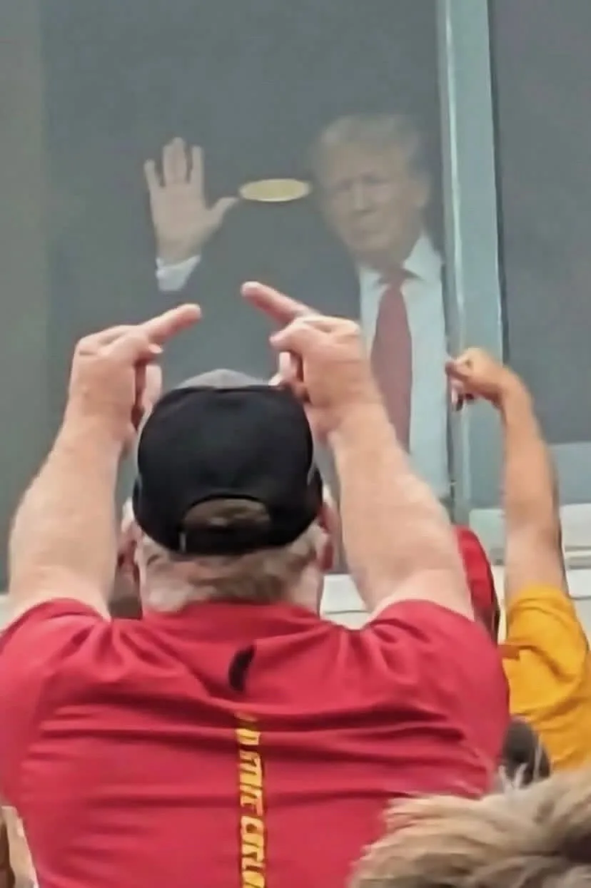 A man in a red shirt and black cap is seen from behind, pointing both index fingers at a blurry image of Donald Trump waving…