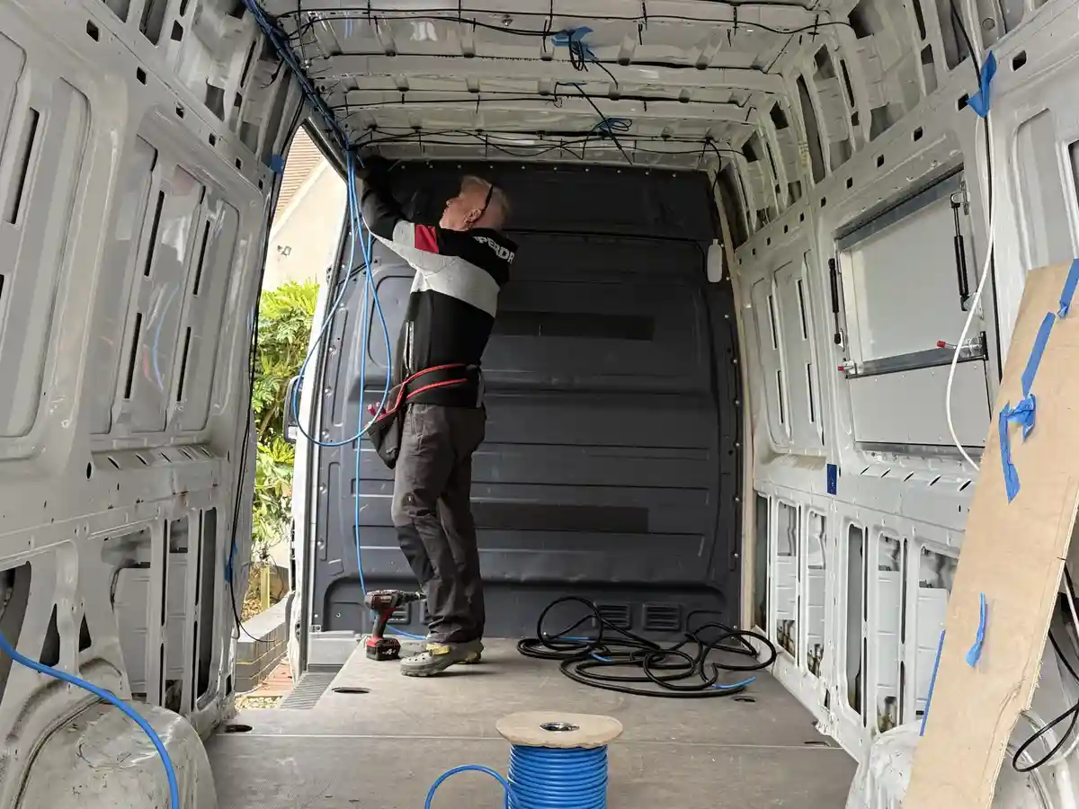 A person is standing inside the bare interior of a van, installing blue electrical wiring along the ceiling. The van's walls…