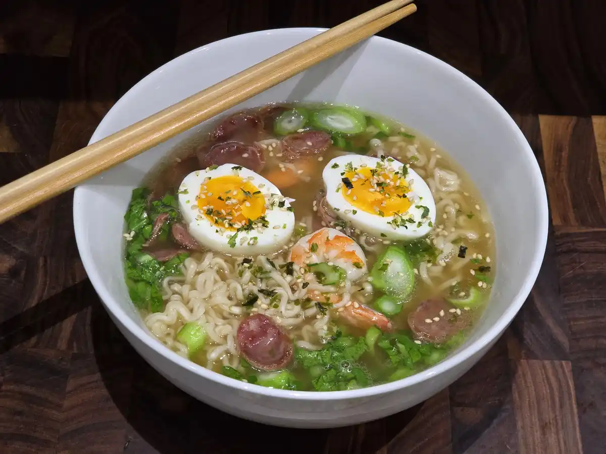 A white bowl of ramen sits on a dark wooden surface, filled with noodles, broth, sliced green onions, shrimp, sausage, and…