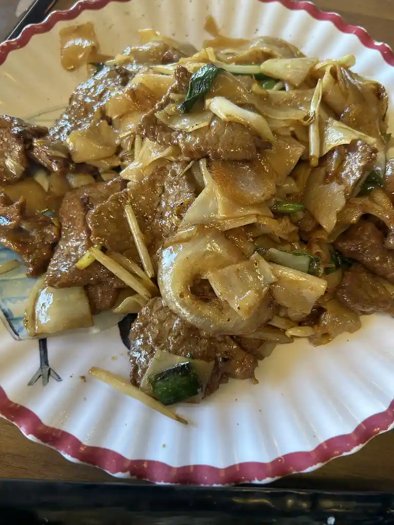 A close-up photo of a plate of stir-fried beef with wide rice noodles, onions, and green onions, served on a white plate…