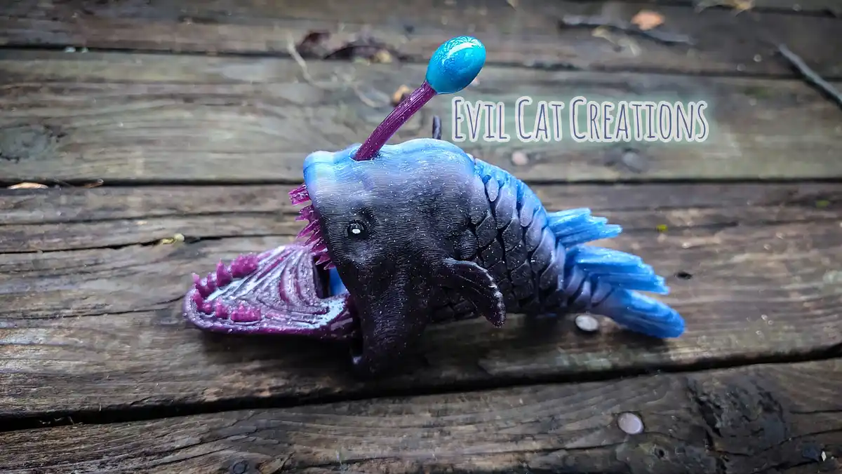 A 3D-printed, stylized anglerfish figurine with a dark blue and purple gradient body, glowing blue lure, and sharp purple…