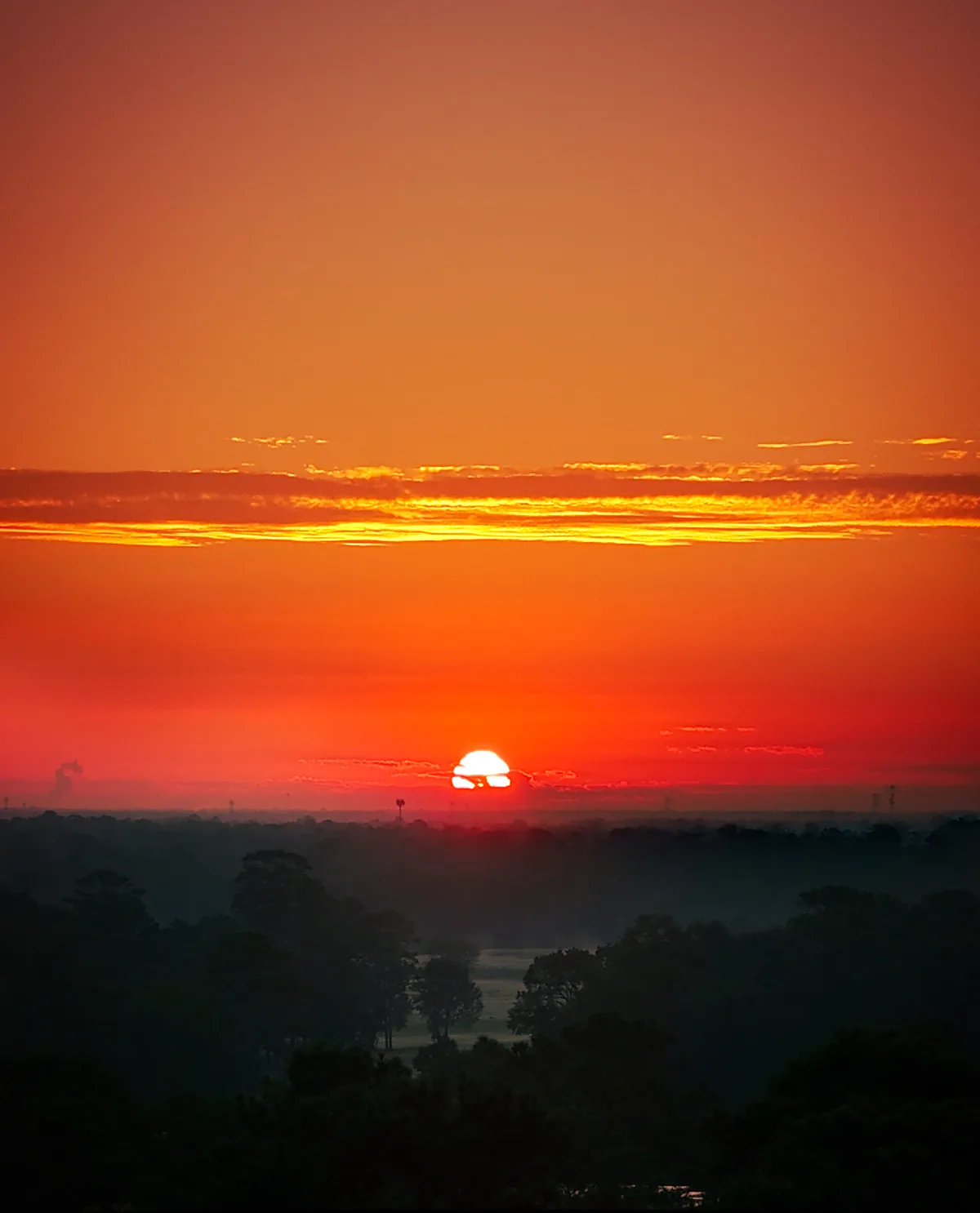A breathtaking sunset over a misty forest, with the sun partially obscured by clouds near the horizon, casting vibrant…