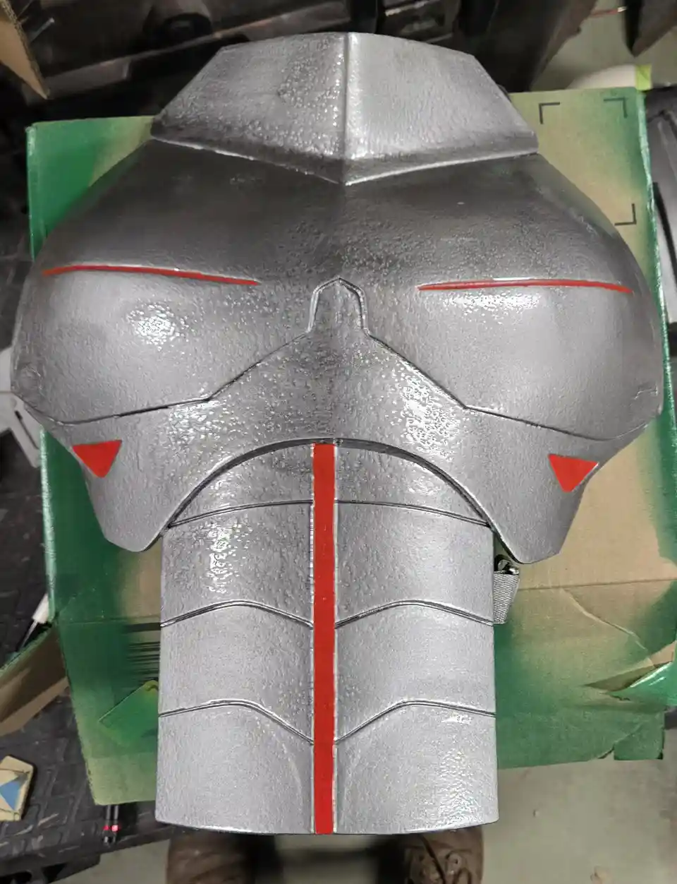 A close-up photo shows a large, silver-painted helmet with red stripes and triangular markings, resting on a green cardboard…