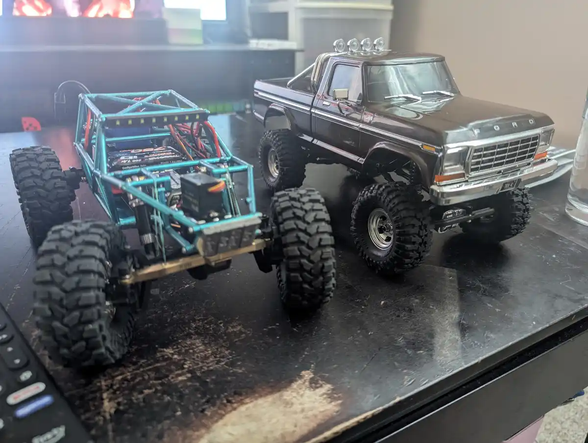 Two remote-controlled monster trucks sit side-by-side on a dark table; the one on the left is an exposed, teal-colored buggy…