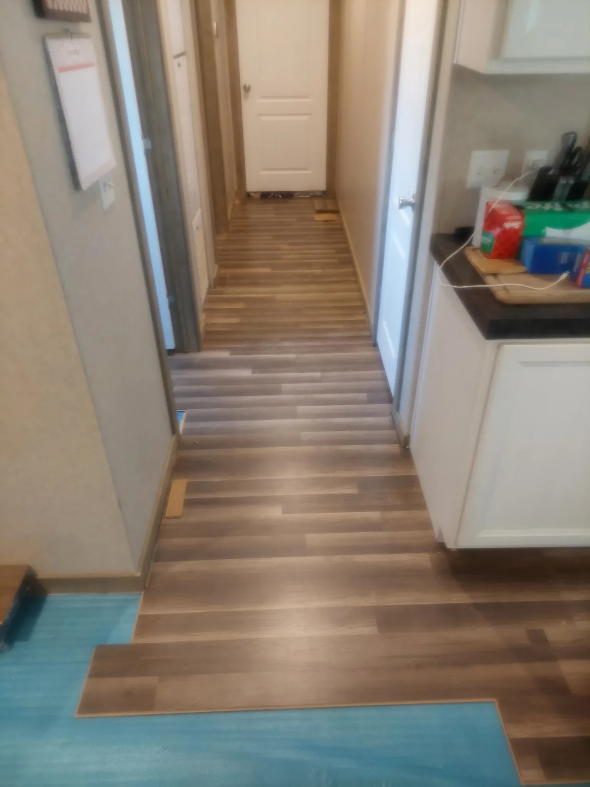 A hallway with newly installed brown wood-look flooring that abruptly ends at a blue carpeted area, with a white door at the…