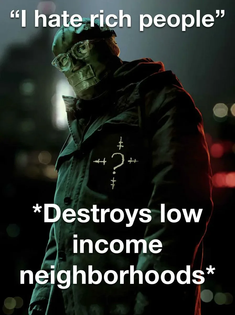 A person wearing a dark coat, glasses, and a taped mouth stands in a dimly lit urban setting with blurred lights in the…