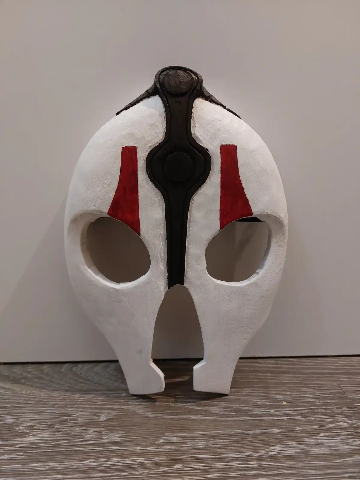 A handmade white mask with red triangular markings on the cheeks and a black central design stands upright on a wooden floor…