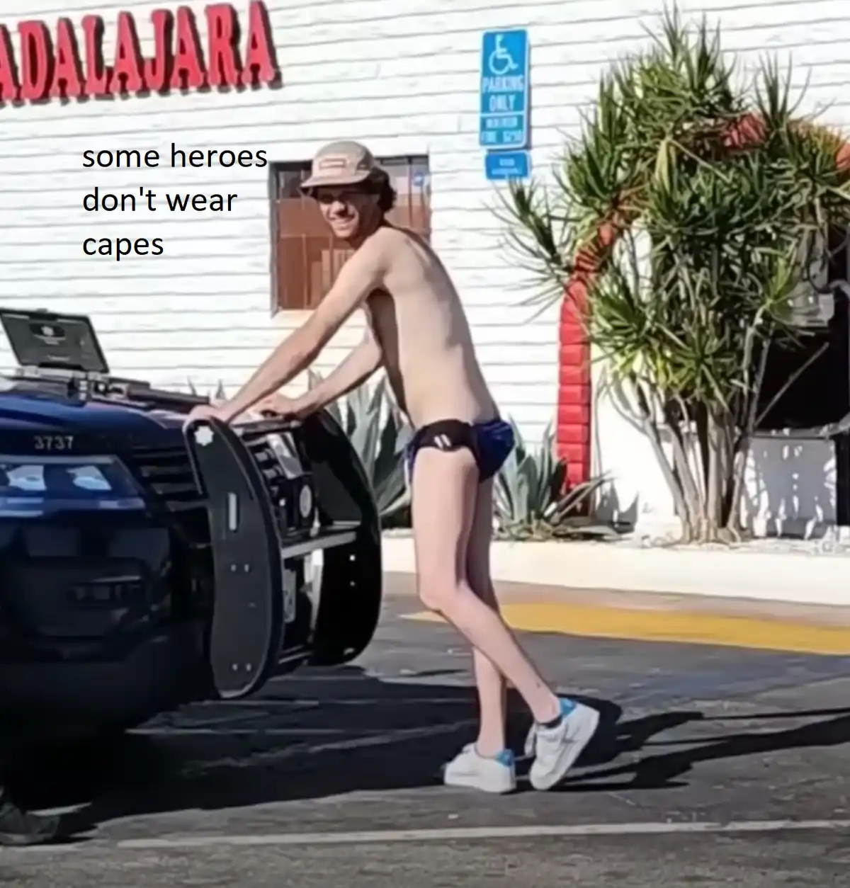 A shirtless young man in blue underwear and a cap is pushing a police car in a parking lot. The car has its hood open and is…