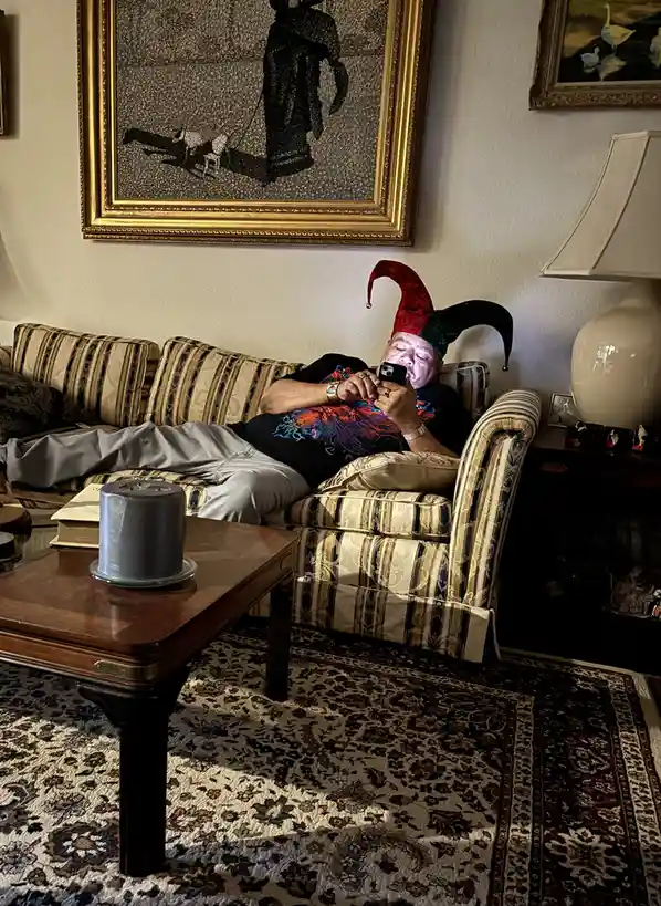 A man is sleeping on a striped couch in a living room, wearing a red and green jester hat while holding a phone. The room…