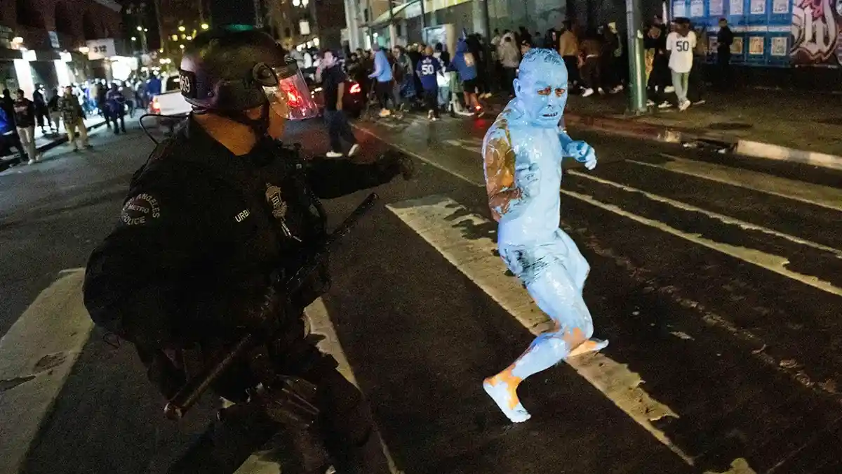 A police officer in full riot gear confronts a person covered in blue paint who is running across a street at night. The…