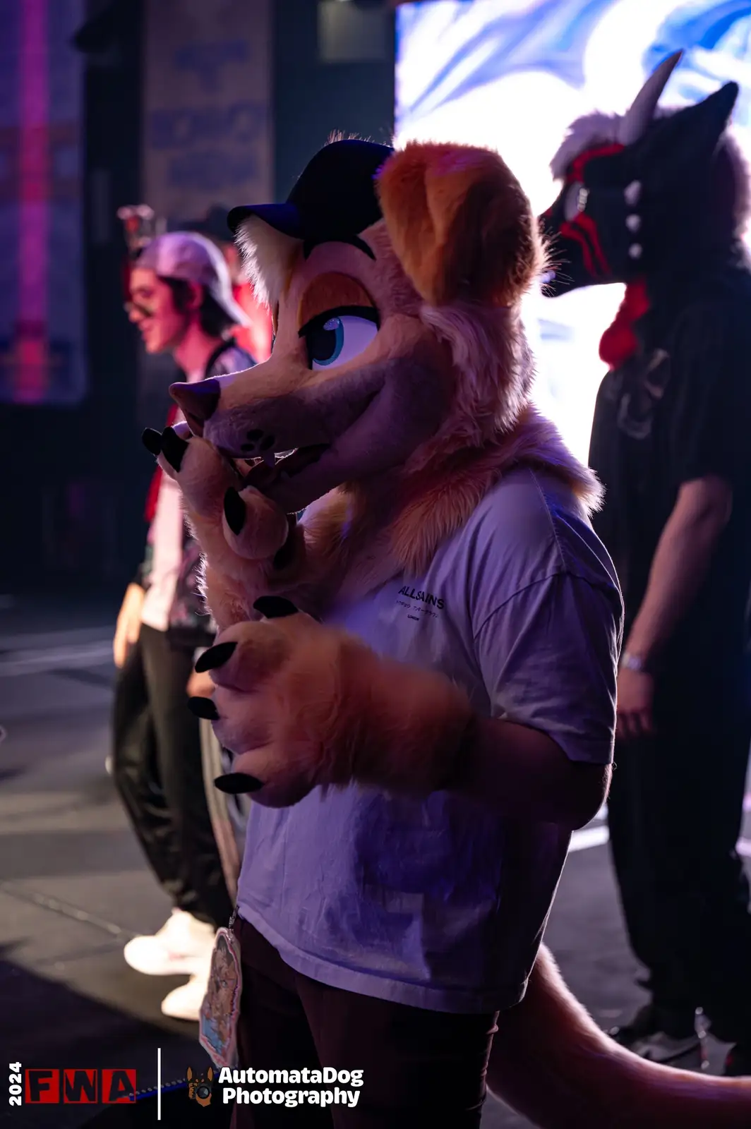 A person in a detailed tan fox fursuit wearing a black cap and a white t-shirt with 'ALLSAINS' printed on it, poses with…