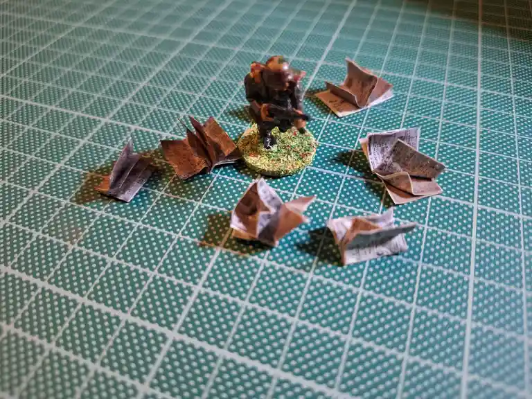A miniature soldier figurine stands on a green grid mat surrounded by several small, folded paper objects resembling books…