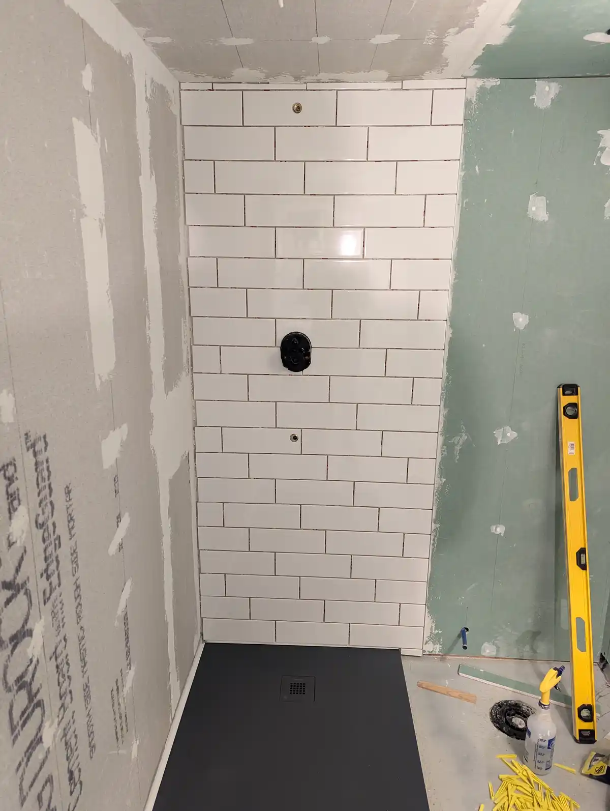 A partially tiled shower stall with white subway tiles on one wall, surrounded by unfinished drywall with visible joint…