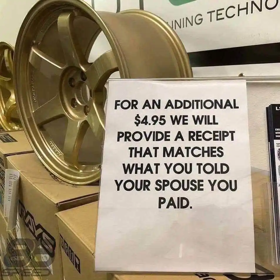 A white paper sign with bold black text is taped to a display stand in a car parts store, positioned in front of…