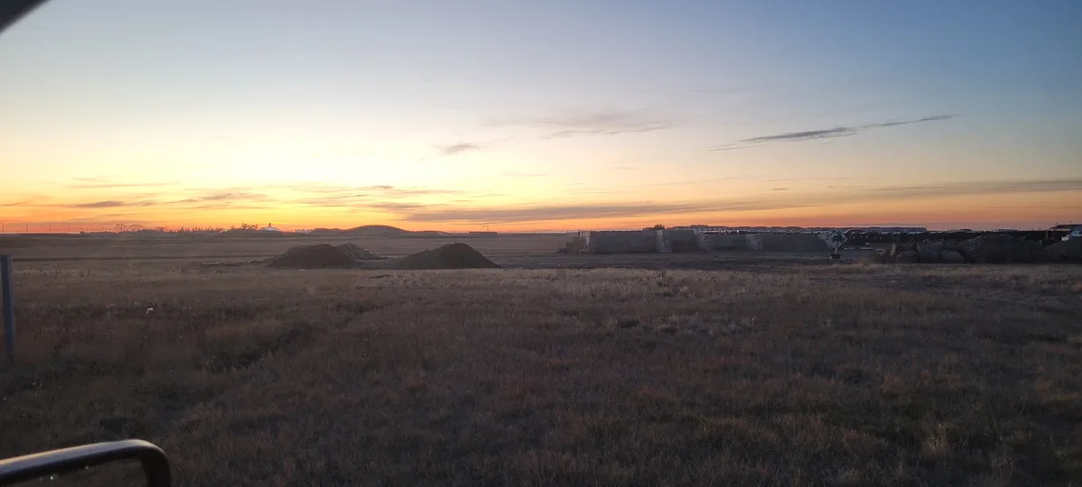 A wide-angle photo captures a vast, flat field at sunset, with dry grass in the foreground, dirt mounds, and a low concrete…