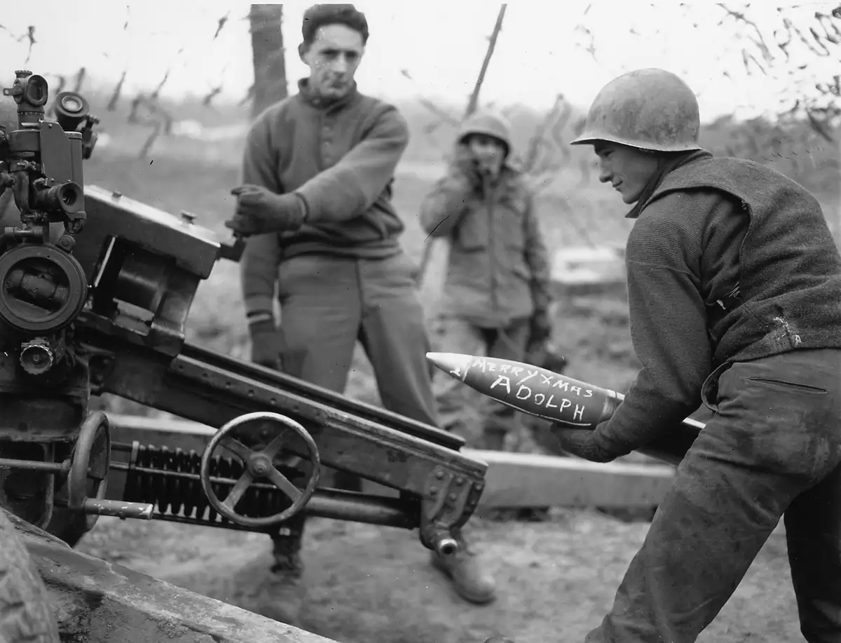 A black-and-white historical photo shows a soldier in a helmet loading a mortar round into a weapon, with two other soldiers…