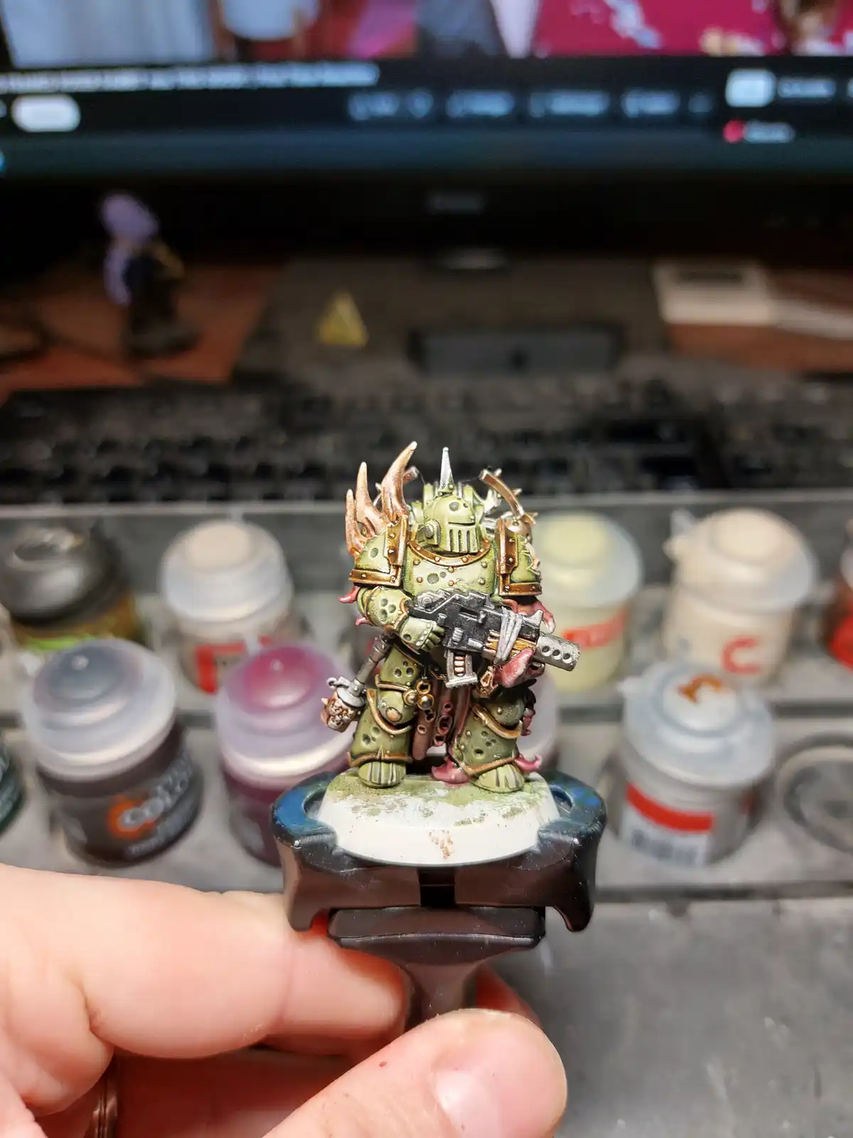 A hand holds a painted green and gold Warhammer 40k miniaturized warrior with horns and a weapon, standing on a base. Behind…