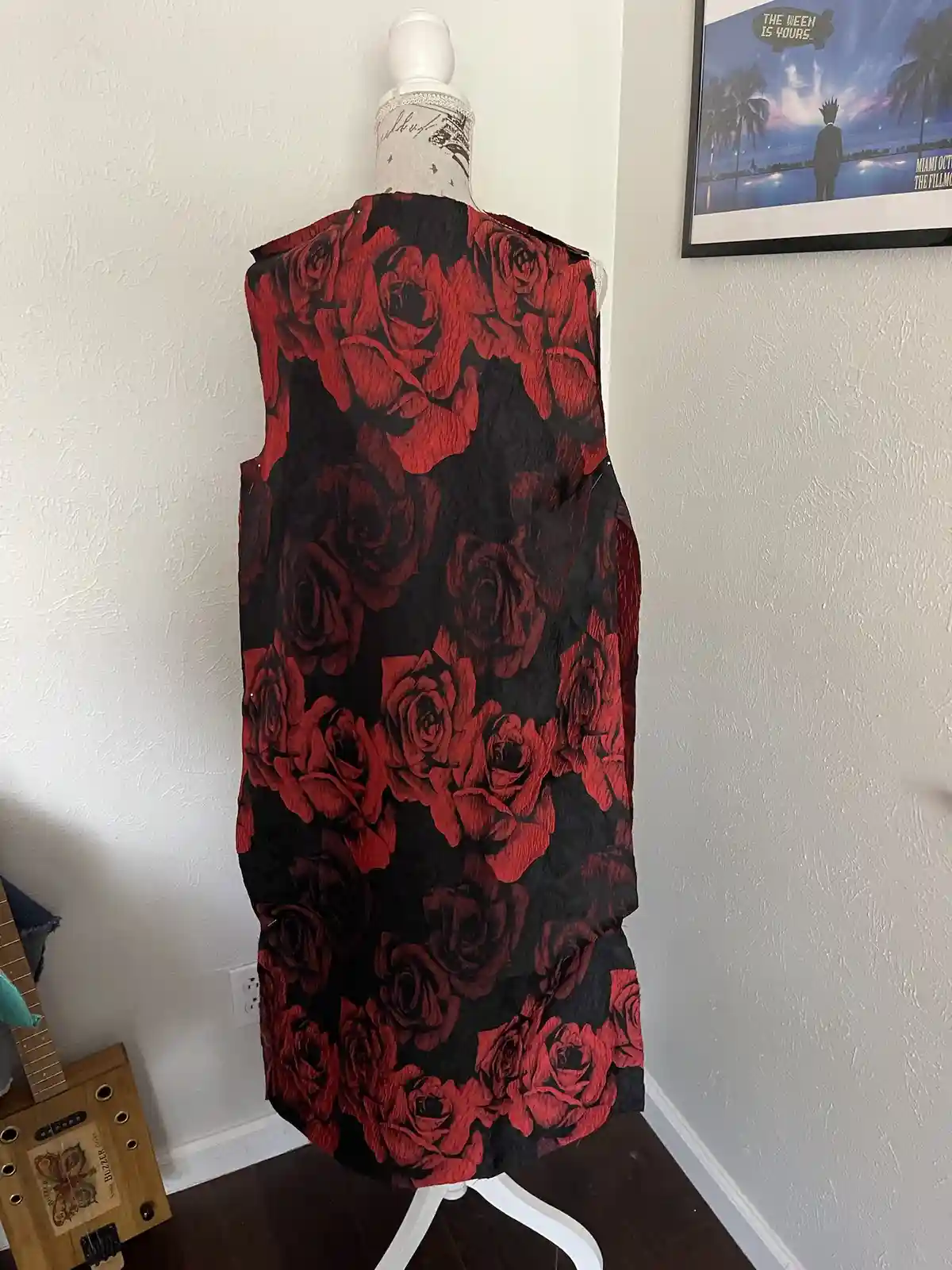 A red and black rose-patterned dress is draped over a white mannequin in a room with a guitar and framed poster on the wall…