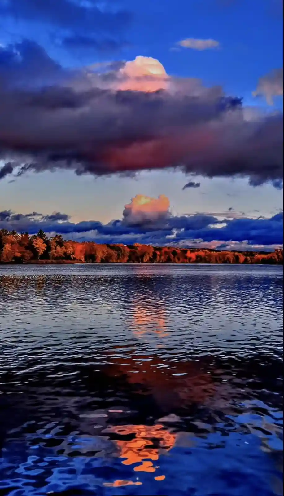A serene landscape photo of a lake at dusk, with vibrant orange and red autumn trees on the far shore and dramatic clouds…