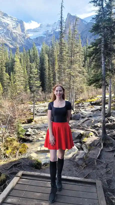 A woman with red hair stands smiling on a wooden boardwalk in a mountainous forest, wearing a black top, a bright red skirt…