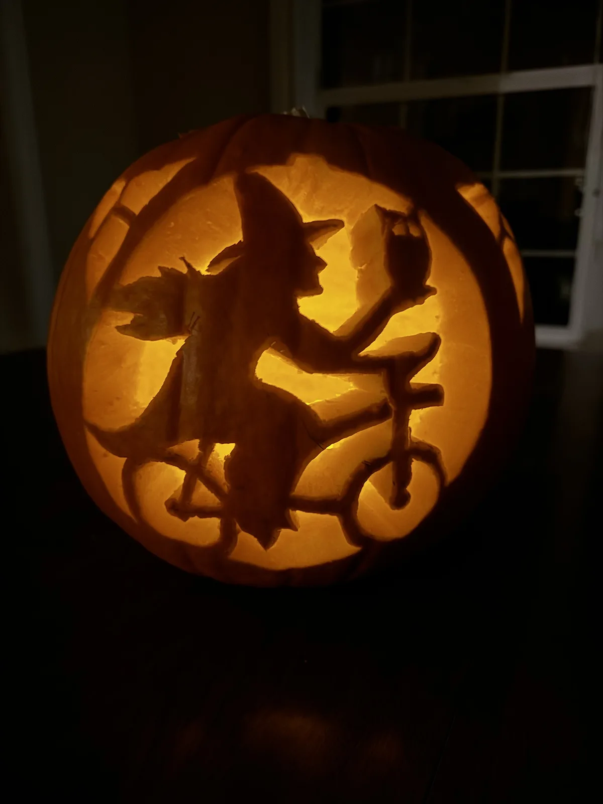 A carved Halloween pumpkin with an internal light source illuminates a silhouette of a witch riding a bicycle, holding a…