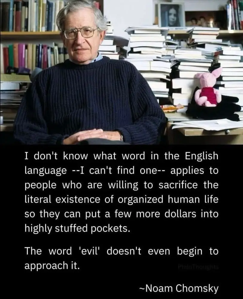 An older man with glasses and a dark sweater sits in a cluttered room filled with stacks of books. A pink stuffed pig sits…