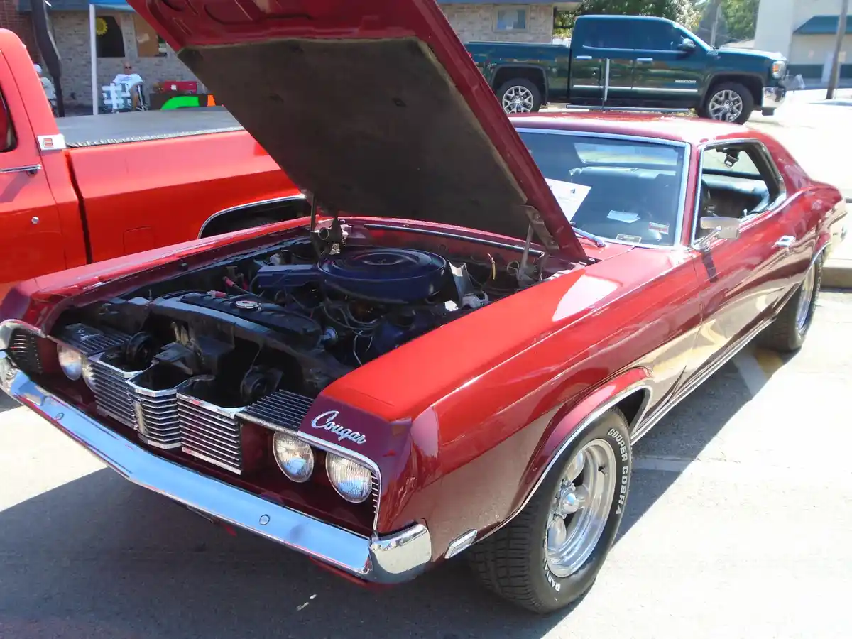 A red classic Ford Cougar muscle car with its hood open, revealing the engine, parked outdoors with other vehicles nearby…