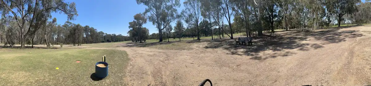 A wide-angle photo of a sunny park with trees, grass, and a dirt path, featuring a blue bucket filled with sand near a disc…