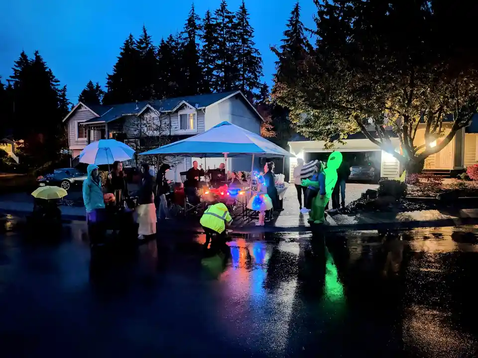 A group of people dressed in Halloween costumes, including a glowing green alien, gather outdoors at night during a…